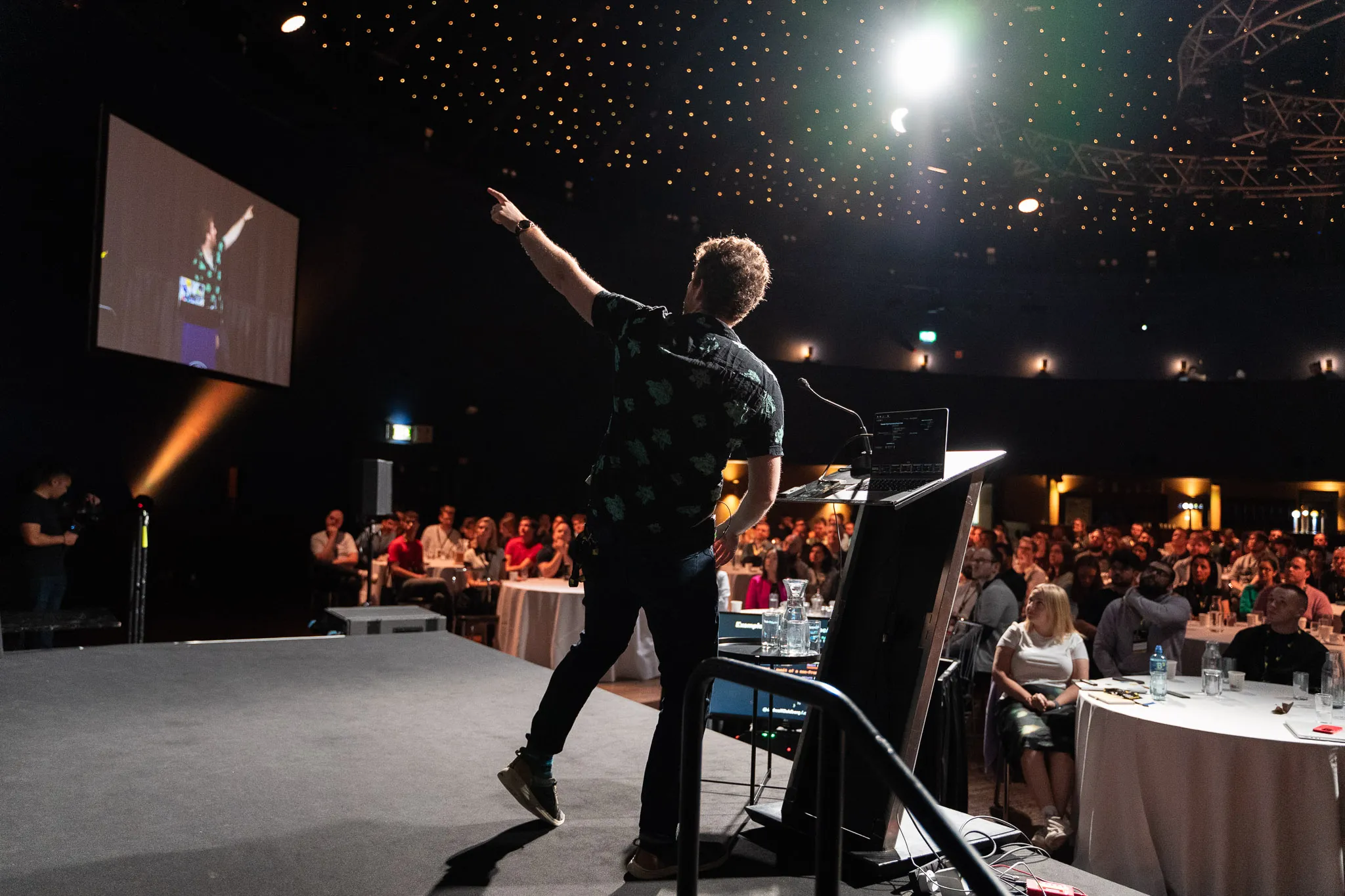 View from behind of me on stage, wearing a black collared shirt with a green Pokemon pattern, pointing to the distance